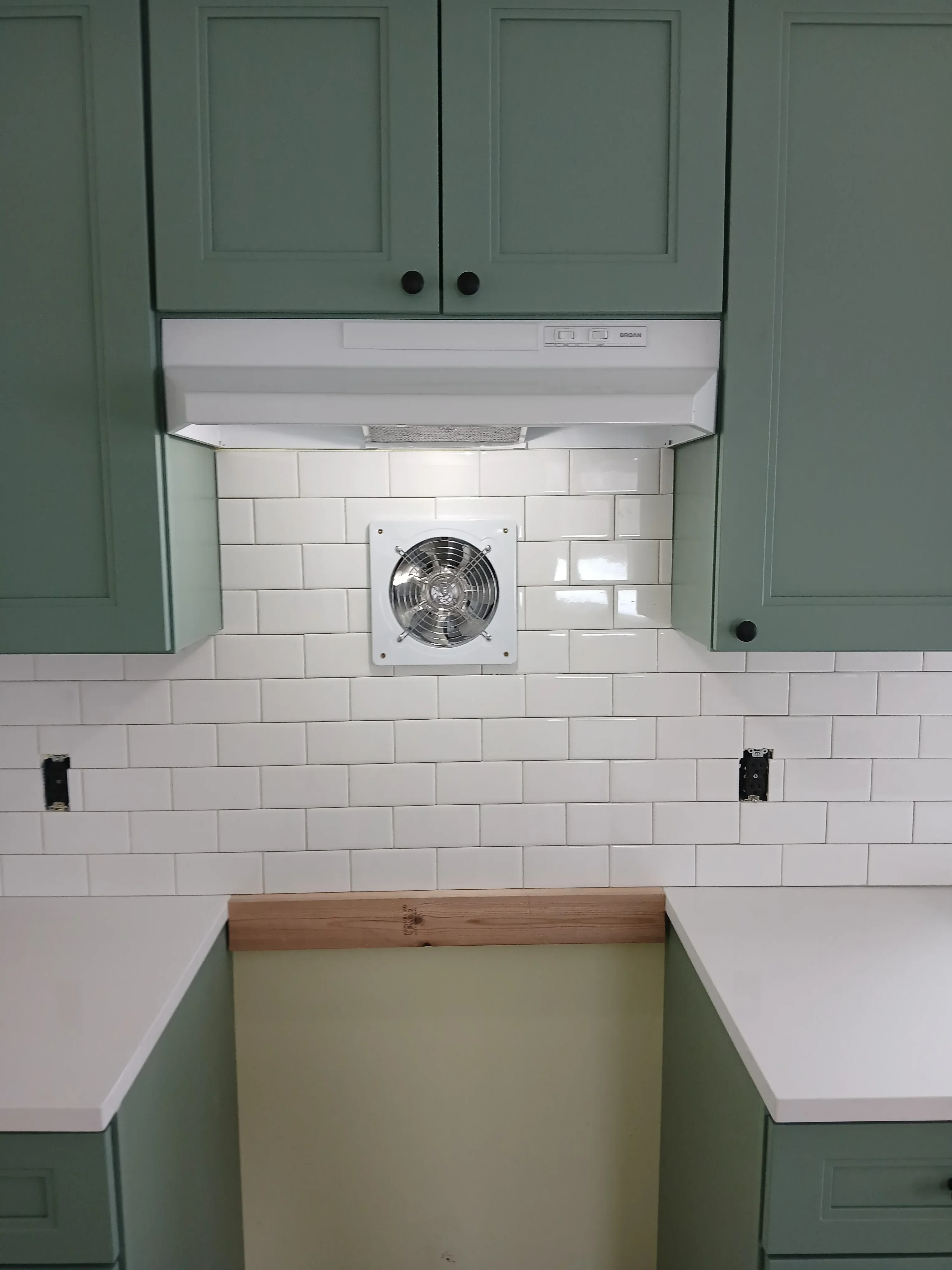 White exhaust fan installed in subway tile backsplash with electrical outlets and green cabinetry